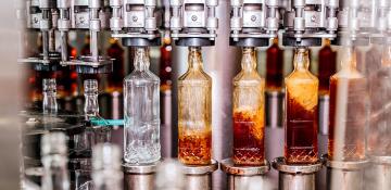 Image shows a bottling line production, with cut glass bottles being filled with a dark liquor. Thr processis automated with the machinery attaching to the neck of the bottle and filling from above.