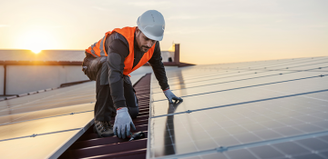 Image shows man wearing white hard hat and high visibility orange dress working on rooftop solar panels. The solar panels are installed on an industrial looking site, on the roof, with the sun setting in the background.